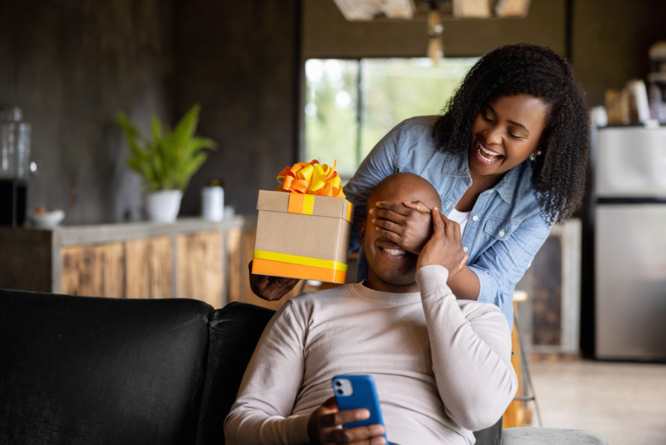 African American woman at home surprising her husband with a gift and covering his eyes